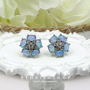 May include: A pair of silver-toned stud earrings. Each earring features a flower design with oval-shaped, light blue opals surrounding a cluster of small, clear gemstones. The earrings are displayed on a white, decorative surface.