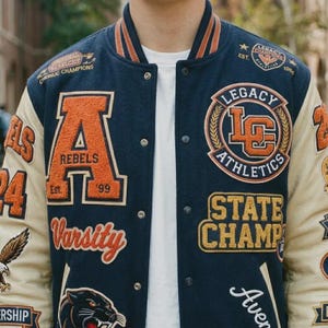 Puede incluir: Una chaqueta universitaria azul marino con mangas color crema, con parches de chenilla naranja y blanco que incluyen una letra A grande, el texto Rebels, Legacy Athletics, State Champ y un gráfico de una cabeza de pantera en el frente.