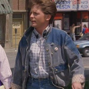 Puede incluir: Una chaqueta vaquera con diseño de patchwork, usada sobre una camisa a cuadros y vaqueros azules. La chaqueta tiene un botón y un pequeño pin. La persona está de pie afuera en una calle de la ciudad.