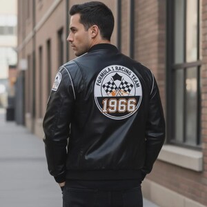 May include: Black leather bomber jacket with white trim and a Formula 1 Racing Team patch. The back features a large circular patch with checkered flags and the year 1966 in orange. The jacket has a classic, sporty design.