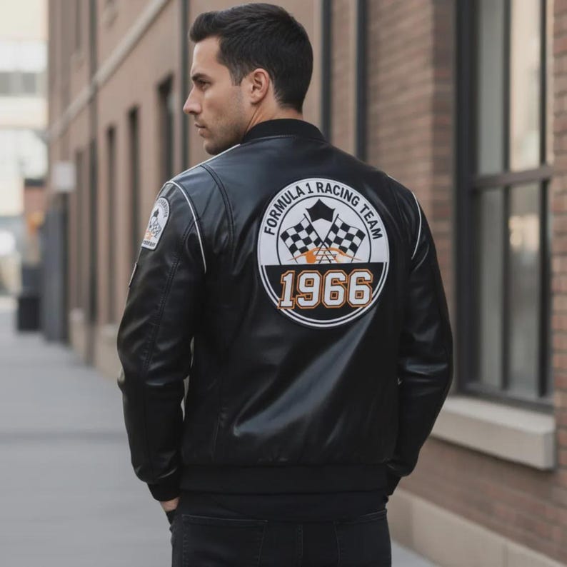 May include: Black leather bomber jacket with white trim and a Formula 1 Racing Team patch. The back features a large circular patch with checkered flags and the year 1966 in orange. The jacket has a classic, sporty design.