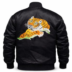 Könnte beinhalten: Schwarze Bomberjacke mit einem gestickten Tiger-Motiv auf dem Rücken. Der Tiger hat orange und gelbe Streifen mit weißen Akzenten. Die Jacke hat einen gerippten Kragen, Bündchen und Saum.