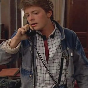 May include: A denim jacket with a light wash over a plaid shirt and a red undershirt. The jacket has a classic design with button closures and pockets. The plaid shirt is white and gray. The person is holding a black telephone.