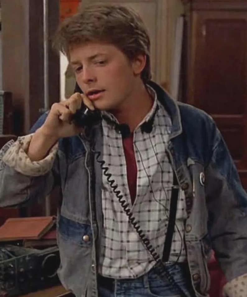 May include: A denim jacket with a light wash over a plaid shirt and a red undershirt. The jacket has a classic design with button closures and pockets. The plaid shirt is white and gray. The person is holding a black telephone.