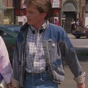 May include: A denim jacket with a light blue wash, featuring a patchwork design and button closures. Worn over a plaid shirt and paired with blue jeans. The jacket has a pin on the lapel and rolled-up sleeves with a patterned lining.
