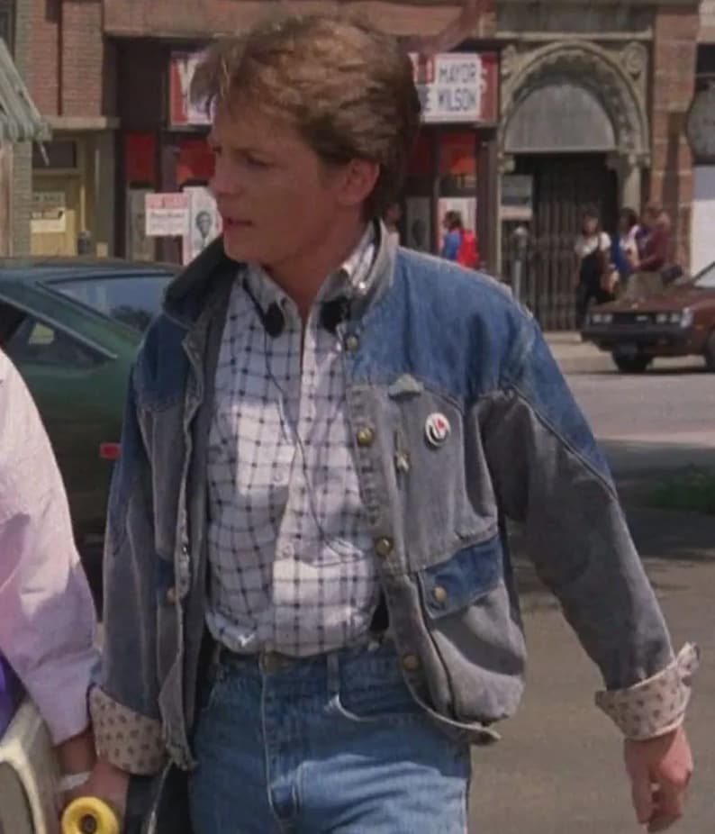 May include: A denim jacket with a light blue wash, featuring a patchwork design and button closures. Worn over a plaid shirt and paired with blue jeans. The jacket has a pin on the lapel and rolled-up sleeves with a patterned lining.