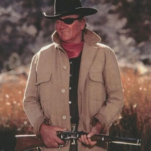 May include: A man wearing a black cowboy hat, eye patch, and tan jacket, holding a rifle. He is also wearing a red scarf and a black shirt. The background is a blurred outdoor scene.