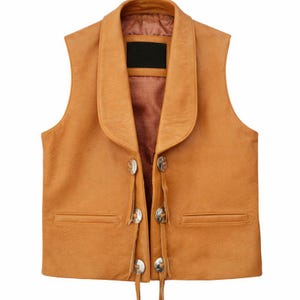 May include: A tan leather vest with a wide collar and silver-coloured buttons. The vest has two front pockets and decorative leather ties. The vest is sleeveless and has a classic Western style.