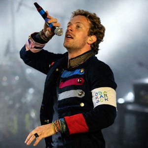 Può includere: Una persona indossa una giacca nera con strisce colorate e bottoni dorati, che tiene in mano un microfono. La giacca ha un bracciale bianco con testo. La persona indossa diversi braccialetti e un anello nero.