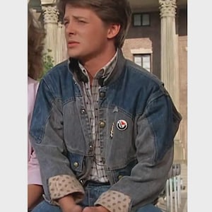 May include: A denim jacket with a two-tone design, featuring a lighter gray on the sleeves and a darker blue on the body. The jacket has a button-up front, a plaid shirt underneath, and a small pin on the chest. The cuffs have a tan and brown polka dot pattern.