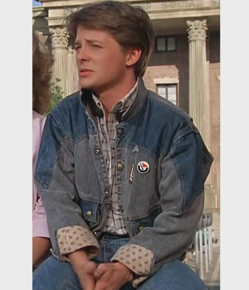 May include: A denim jacket with a two-tone design, featuring a lighter gray on the sleeves and a darker blue on the body. The jacket has a button-up front, a plaid shirt underneath, and a small pin on the chest. The cuffs have a tan and brown polka dot pattern.