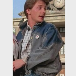 May include: A denim jacket with a faded blue wash and a button-down plaid shirt underneath. The jacket has a button closure and a small pin on the chest. The person is wearing blue jeans.