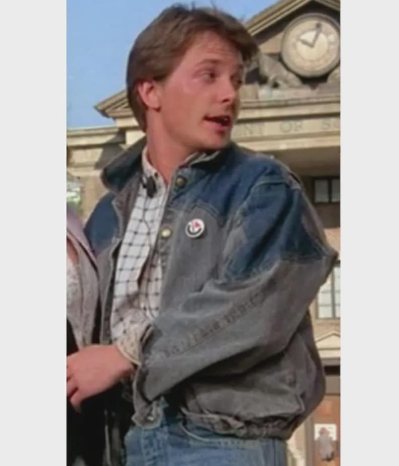 May include: A denim jacket with a faded blue wash and a button-down plaid shirt underneath. The jacket has a button closure and a small pin on the chest. The person is wearing blue jeans.