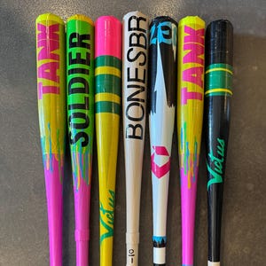 May include: A collection of colorful baseball bats. The bats feature vibrant designs with text like "TANK", "SOLDIER", and "BONESBR". Each bat has a unique color scheme, including pink, green, yellow, and black, with contrasting accents. The bats are arranged vertically.
