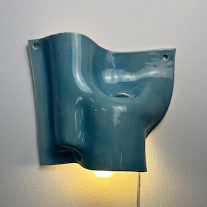 May include: A wall-mounted light fitting with a glossy, blue, undulating ceramic shade. The sculptural form features soft curves and two small circular holes for mounting. A warm, glowing light bulb is visible at the bottom, with a thin power cord.
