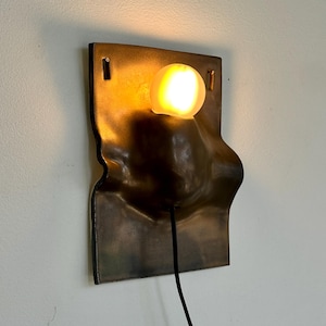 May include: A bronze-colored wall sconce with a unique, textured design. The fixture features a single, illuminated bulb, casting a warm glow. A black cord extends from the sconce. The sconce is mounted on a white wall.