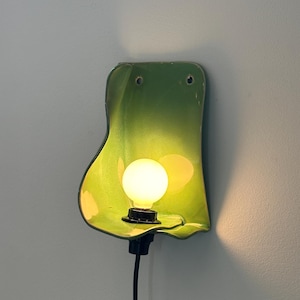 May include: A wall-mounted lamp featuring a glossy, lime green, organic-shaped ceramic shade with a curved, flowing silhouette. A single round, warm-glowing light bulb is centred within the shade, connected to a black socket and a black power cord.