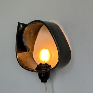 May include: A wall-mounted light fixture with a black, curved metal shade and a warm, glowing lightbulb. The interior of the shade is gold-toned, creating a contrast with the dark exterior. A black socket and clear cord complete the design.