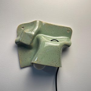 Może przedstawiać: Ścienna lampa ceramiczna w kolorze jasnej miętowej zieleni z ciemnymi nakrapianymi wzorami. Przedmiot ma nieregularny, pofałdowany i organiczny kształt z dwoma otworami montażowymi w górnych rogach oraz pojedynczą białą żarówką skierowaną w dół.