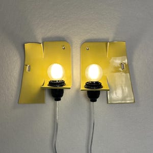 May include: Two yellow ceramic wall sconces with an irregular, folded shape. Each fixture features a black socket holding a glowing, round white light bulb. A thin, silver-colored electrical cord hangs downward from the base of each lamp.