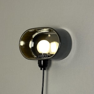 K&ouml;nnte beinhalten: Eine moderne Wandleuchte mit dunkelgrauer Au&szlig;enseite und goldfarbenem Innenraum. Die Leuchte hat eine ovale Form mit einer Gl&uuml;hbirne im Inneren. Ein schwarzes Kabel erstreckt sich von der Basis.