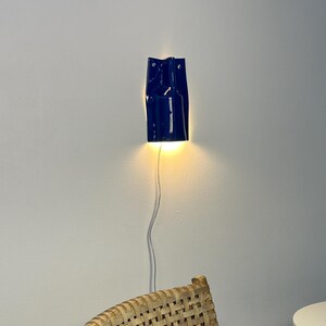 Puede incluir: Un aplique de pared de cer&aacute;mica azul oscuro brillante con un dise&ntilde;o vertical y plegado. La l&aacute;mpara est&aacute; montada en una pared y emite una luz c&aacute;lida dirigida hacia abajo. Un cable de alimentaci&oacute;n blanco y fino cuelga desde la base.