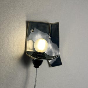 May include: A ceramic wall sconce with a dark blue glaze. The sconce has a unique, folded design with a small shelf to hold a round, glowing light bulb. A black socket and a gray cord complete the fixture.