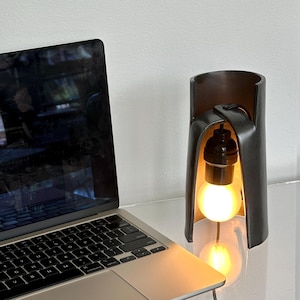 May include: A modern, dark gray desk lamp with a curved design, featuring a visible lightbulb. The lamp is placed on a clear surface next to a laptop computer. The lamp's design is minimalist and industrial.