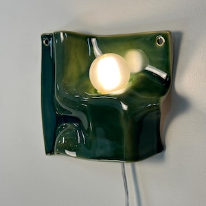 May include: A wall-mounted ceramic light fixture with a glossy, deep green, undulating surface. The piece features a circular, glowing white bulb integrated into the center of the curved form, with two small holes at the top corners for mounting.