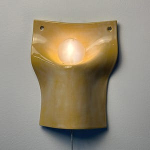 May include: A wall-mounted ceramic sconce with a glossy yellow finish and a curved, concave shape. The fixture features two circular mounting holes at the top corners and a central, glowing white spherical light bulb extending from the middle.