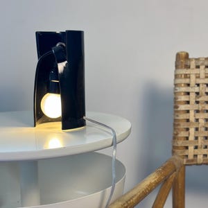 May include: A black cylindrical table lamp with a glossy finish and an open front section. The lamp features a single exposed light bulb emitting a warm glow, connected to a thin, clear power cord that extends from the base of the fixture.