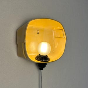 May include: A wall-mounted lamp featuring a bright yellow, rounded rectangular shade with a smooth finish. A small, frosted spherical light bulb is centered within the shade, attached to a black socket base with a thin, light-colored power cord.