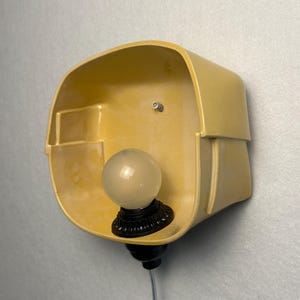 May include: A wall-mounted lamp featuring a smooth, yellow ceramic shade with an asymmetrical, rounded rectangular shape. A black socket holds a single, frosted white globe light bulb, and a thin gray power cord extends from the bottom.