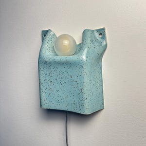 May include: A wall-mounted light fixture made of light blue ceramic with a dark brown speckled finish. The rectangular, folded-form shade features two top mounting holes and holds a single round, frosted white light bulb in the center.