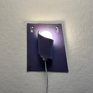 Pode incluir: Uma arandela de parede moderna com uma placa traseira retangular roxa escura. Uma sombra curva, roxa escura, abriga uma l&acirc;mpada redonda, branca e brilhante. A arandela &eacute; conectada com um fio cinza fino.