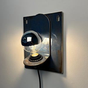 May include: A wall-mounted lamp featuring a rectangular, dark blue ceramic backplate with a curved shelf holding a light bulb. The bulb has a reflective silver top half and a clear bottom half, emitting a warm glow from a black socket base.