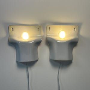 May include: A pair of white wall-mounted sconces with a smooth, curved, T-shaped design. Each fixture features a central circular light source emitting a warm glow, with two small rectangular mounting holes at the top and a thin power cord extending downward.