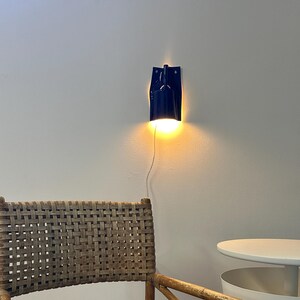 Puede incluir: Un sill&oacute;n de rat&aacute;n trenzado con acabado en marr&oacute;n claro natural y estructura de refuerzo en cruz. Sobre la silla, una l&aacute;mpara de pared azul oscuro emite una luz c&aacute;lida y brillante. A la derecha, una mesa auxiliar redonda blanca con estante.