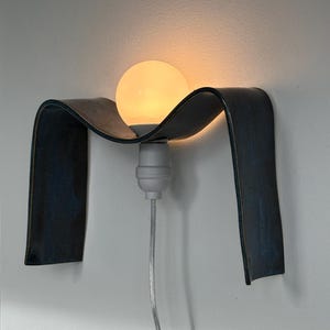 May include: A modern wall sconce featuring a dark blue, curved, ribbon-like design. The sconce holds a round, glowing light bulb. The light fixture is mounted on a white wall, with a gray cord extending downwards.