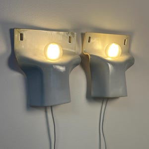 May include: A pair of wall-mounted light fixtures featuring a smooth, pale blue ceramic body with a curved, sculptural form. Each fixture holds a single glowing, spherical light bulb and includes two rectangular mounting holes at the top.