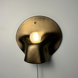 May include: A wall-mounted lamp featuring a circular, metallic bronze-toned shade with a fluid, organic, and concave shape. A single, small, spherical frosted light bulb is positioned in the center, emitting a warm glow. A thin, clear power cord hangs below.