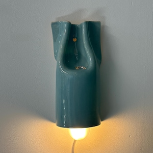 May include: A wall-mounted ceramic light fixture with a glossy teal finish and a sculptural, folded form. The fixture features a central opening and emits a warm, downward-facing glow from a light bulb positioned at the base, with a thin power cord.