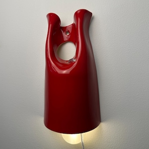 May include: A glossy red wall-mounted lamp with a smooth, curved, elongated form featuring a central circular cutout near the top. The fixture has a wide, open base that emits a warm, soft light from a single bulb, with a clear power cord extending downward.