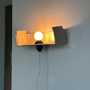 May include: A modern wall sconce with a warm, glowing light bulb. The sconce has a beige, speckled shade and a black bulb socket. The light is mounted on a white wall, with a visible power cord.
