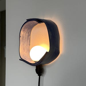 Puede incluir: Una l&aacute;mpara de pared moderna con una pantalla curva azul oscuro. La pantalla rodea una bombilla redonda y blanca. Un cable negro se extiende hacia abajo desde la l&aacute;mpara.
