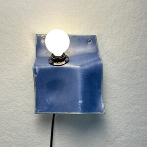 May include: A wall-mounted light fixture featuring a square, blue ceramic panel with a curved, folded shape. A single round, glowing white light bulb is mounted in the upper left section of the ceramic piece, which includes two small metal eyelets.
