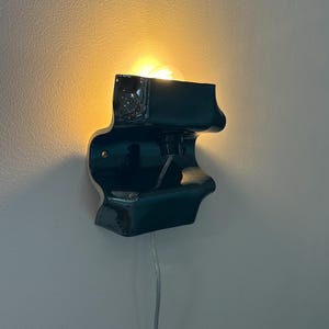 May include: A dark teal ceramic wall sconce with a glossy finish and an undulating, organic shape. The fixture features a central light bulb socket emitting a warm glow, with a clear power cord extending downward from the base of the unit.