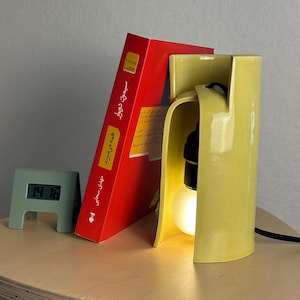 May include: A glossy yellow ceramic table lamp with a curved, open-front design housing a single light bulb. Next to it stands a red book with Persian text on the spine and a small, light green digital clock displaying the time 14:16.
