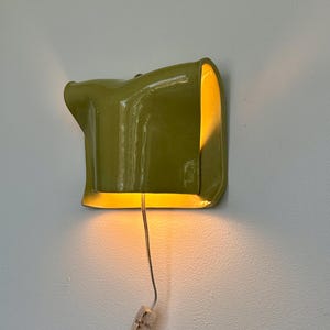 May include: A modern, olive green wall sconce with a unique, curved design. The sconce emits a warm, yellow light, creating a soft glow. A silver wire and a clear connector are visible. The sconce is mounted on a white wall.
