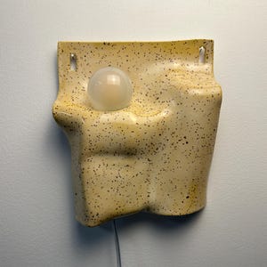 May include: A wall-mounted ceramic light fixture with a speckled cream and yellow glaze. The sculptural form features an irregular, organic shape with a recessed circular opening holding a single frosted glass globe bulb and two top mounting holes.